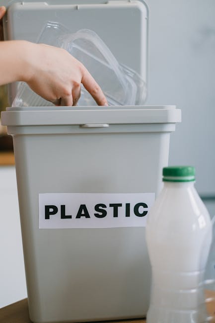 A grey plastic recycling bin with a black and white label reading 'PLASTIC' is open, revealing transparent plastic packaging inside. Next to the bin, on a wooden surface, there is a partially visible